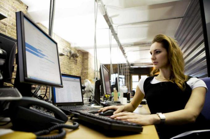 Ženska, ki sedi za mizo in uporablja prenosni računalnik z zunanjo tipkovnico in monitorjem.