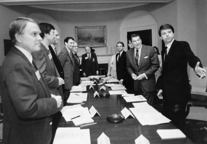 Črno -bela podoba Ronalda Reagana in več drugih moških v oblekah okoli dolge konferenčne mize